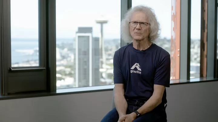 A man with grey hair and glasses in front of a windows that looks onto Seattle's Space Needle. 