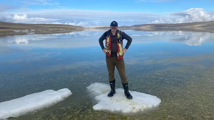 A man wearing glasses, a Vancouver Canucks baseball cap and rubber boots standing on an ice patch in the middle of a body of water.