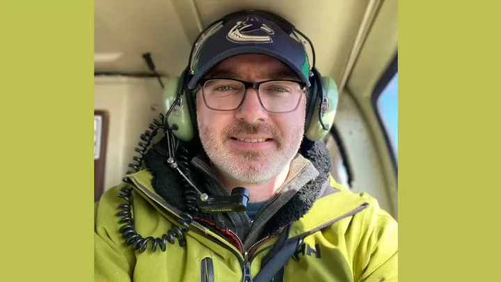 A man wearing glasses and a Vancouver Canucks baseball hat, headphones and microphone inside the cockpit of a helicopter.