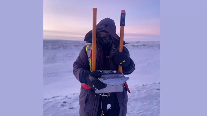 A person wearing a parka and holding two poles and survey equipment with a snowy landscape behind them.