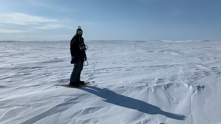 A person wearing a parka, sunglasses and snowshoes holding survey equipment in front of a snowy backdrop.