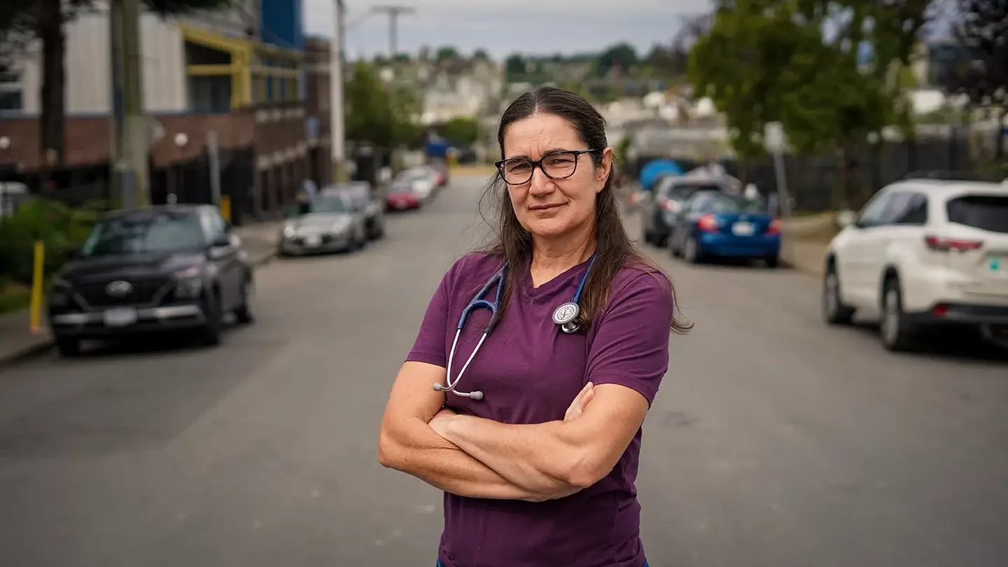 A woman wearing glasses and a stethoscope around her neck standing with her arms crossed in the middle of a road.