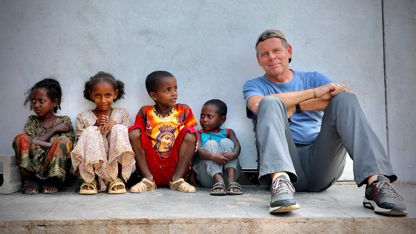 A man wearing a backwards baseball cap sits beside four young children against a wall. 