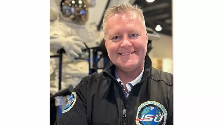 Man smiling and wearing a flight jacket with a badge that reads ISU: International Space University in front of a life-size model of an astronaut.