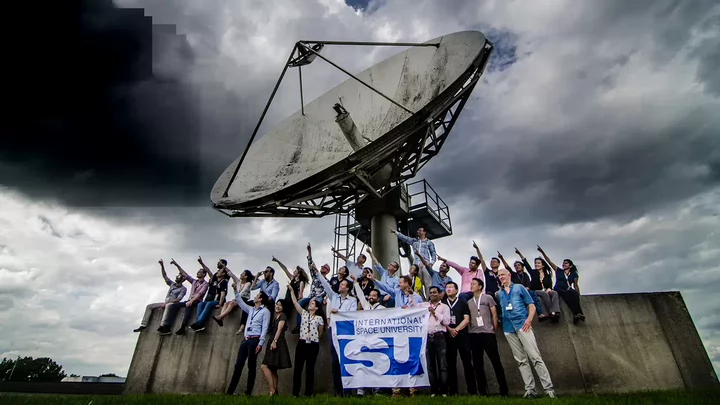 Group of people pointing at the sky while standing in front of a satellite dish and holding a banner that says International Space University.