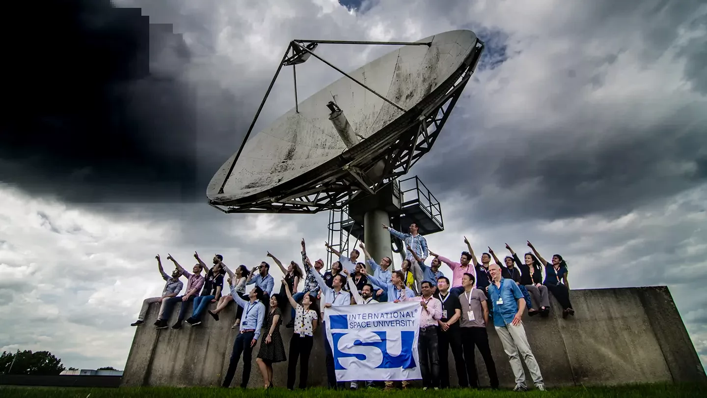 Group of people pointing at the sky while standing in front of a satellite dish and holding a banner that says International Space University.
