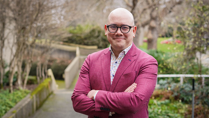 Man wearing glasses, pink blazer and polka dot shirt, standing cross armed outdoors.
