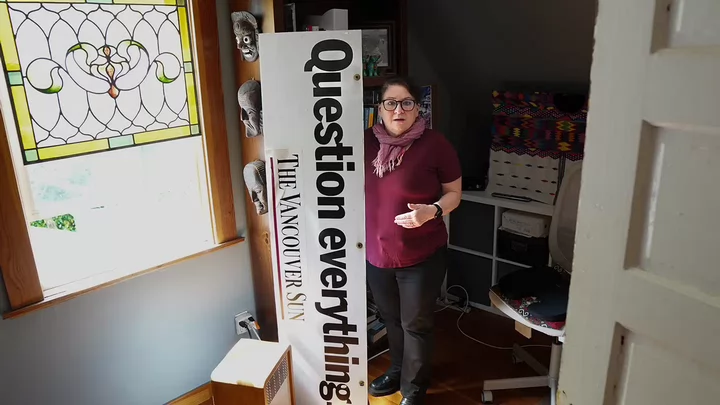 A woman standing in a home office next to a stained glass window and holding a sign that says Vancouver Sun: Question Everything.
