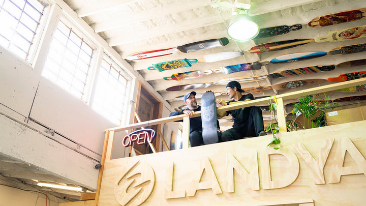 Two people sitting in a loft space with a neon ‘Open sign’ and skateboards lining the ceiling.