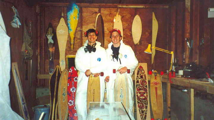 Two people in white jumpsuits posing around various unfinished skateboards in a wooden work shed.