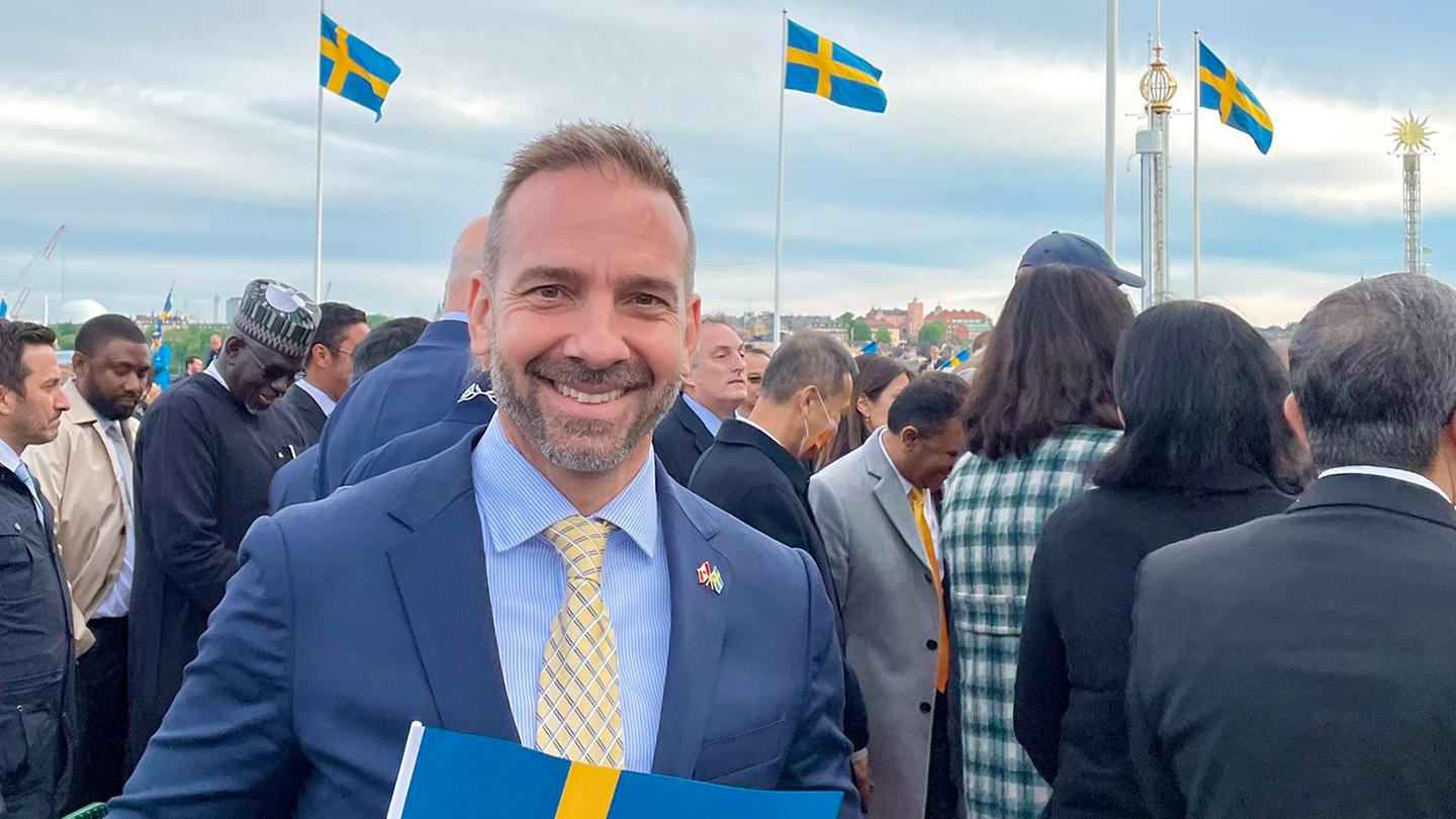 Man in blue suit at public event with Swedish flags in the background. 