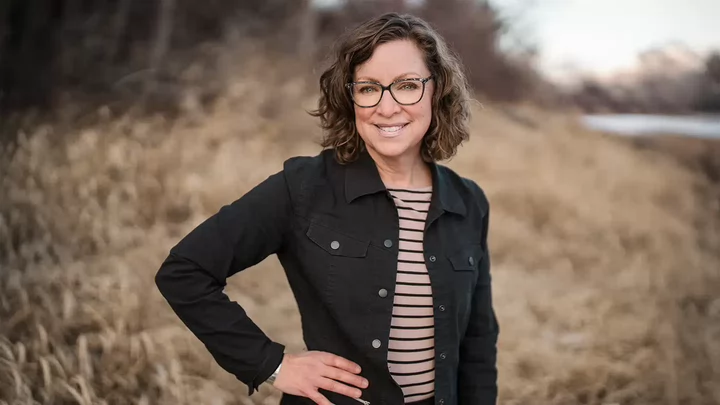 A woman with glasses smiling and standing with her hand on her hip outdoors.