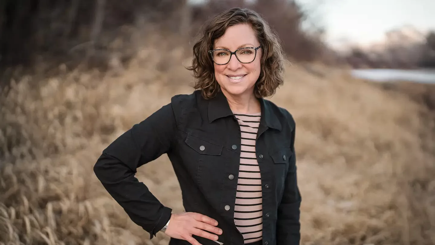 A woman with glasses smiling and standing with her hand on her hip outdoors.