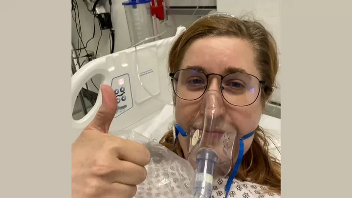  A woman with glasses in a hospital bed wearing an oxygen mask and giving the thumbs up.