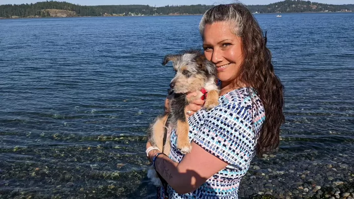 A woman holding a small dog and smiling while standing on a beach. 