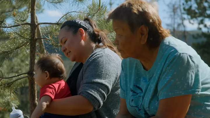 A woman holding a toddler sits beside another woman surrounded by trees.