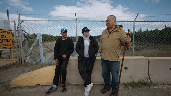 Three people stand in front of a chainlink fence with barbed wire at the top.