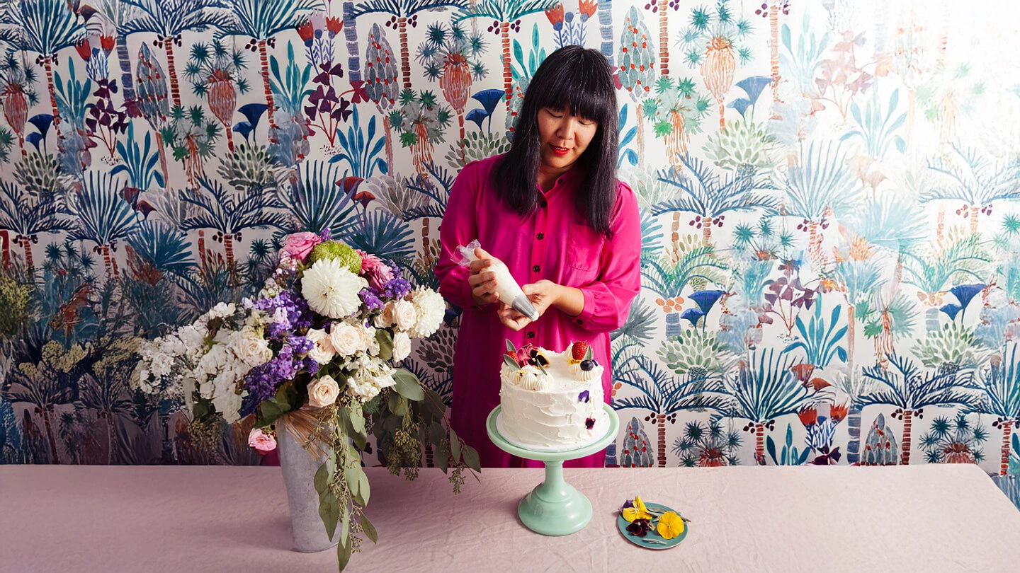 A woman decorating a cake beside a vase of flowers and flowery wallpaper behind her.