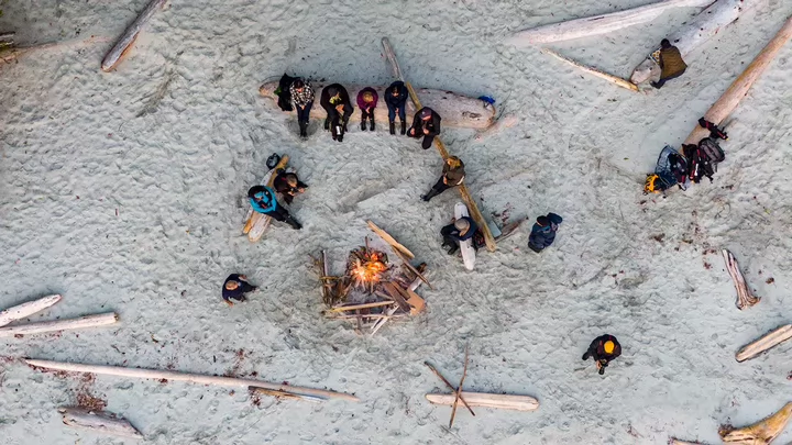 Overhead shot taken by a drone of several people sitting on logs gathered around a campfire on a beach. 