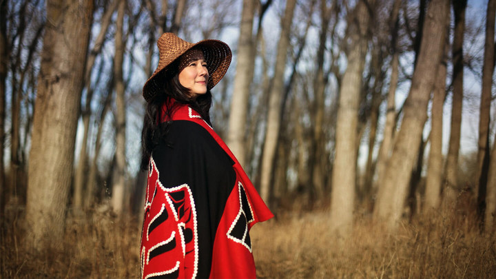 Woman standing in the woods wearing Indigenous blanket and hat. 