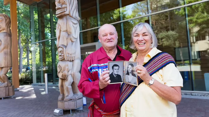 An older man and woman hold up younger photos of themselves while standing in front of a building with Indigenous totem poles.  
