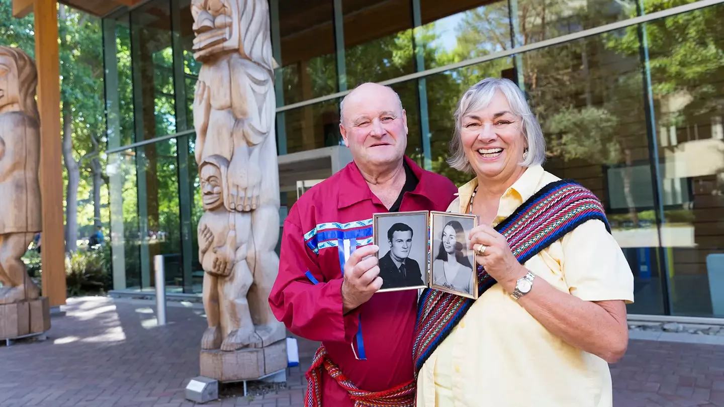 An older man and woman hold up younger photos of themselves while standing in front of a building with Indigenous totem poles.  