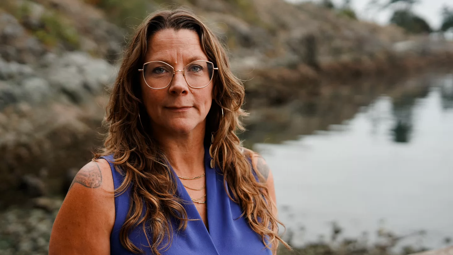A woman with long hair and glasses standing in front of a beach.
