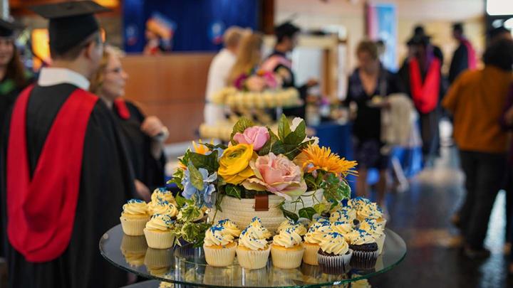 A plate of cupcakes and flowers in foreground with guests at a convocation party in background