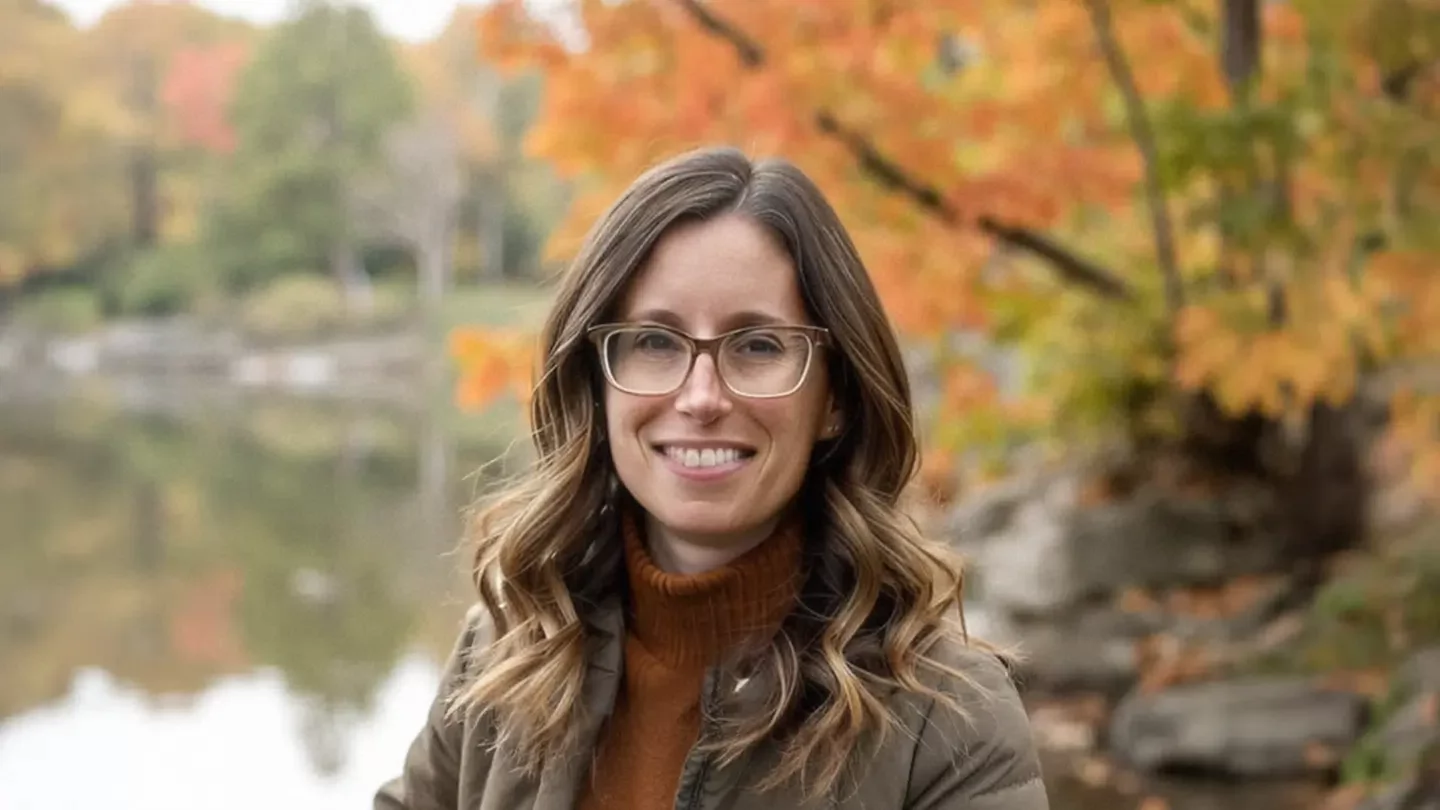 A woman with long hair and glasses smiling while standing in front of a body of water reflecting leafy trees.