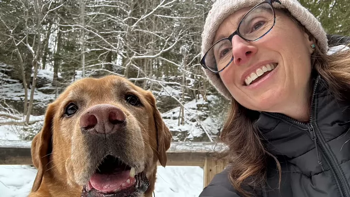 A woman wearing glasses and a toque on a wintry day smiling and taking a selfie with her dog. 