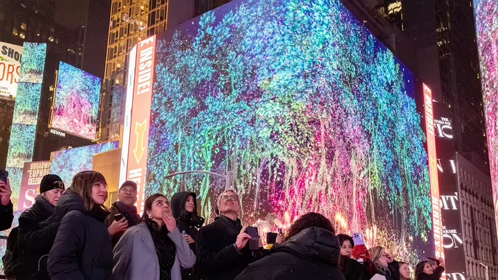 A group of people standing on a street corner looking up at a digital image of a tree on a digital billboard in New York City's Times Square.