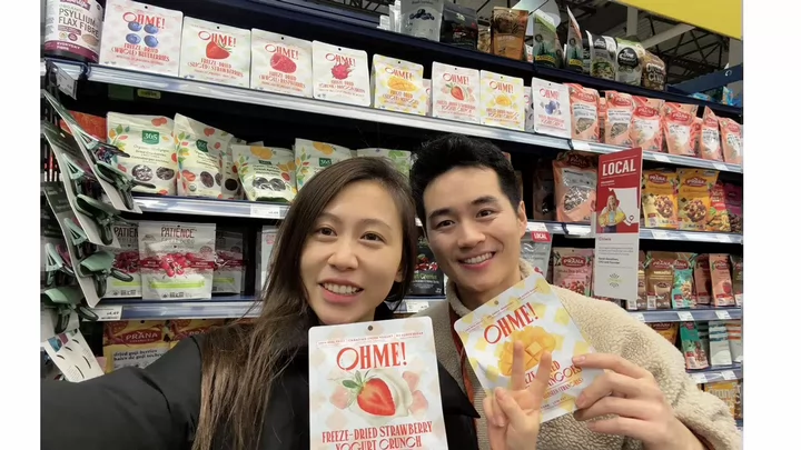 Two people taking a selfie in a grocery store holding up packages of OHME! freeze dried fruit.