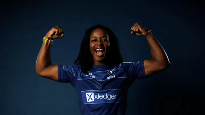 A woman's rugby player raises her arms in the air like she's flexing her muscles while smiling.
