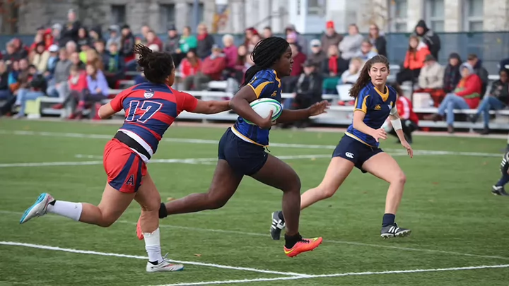 A woman's rugby player holding the ball runs past her opponent.