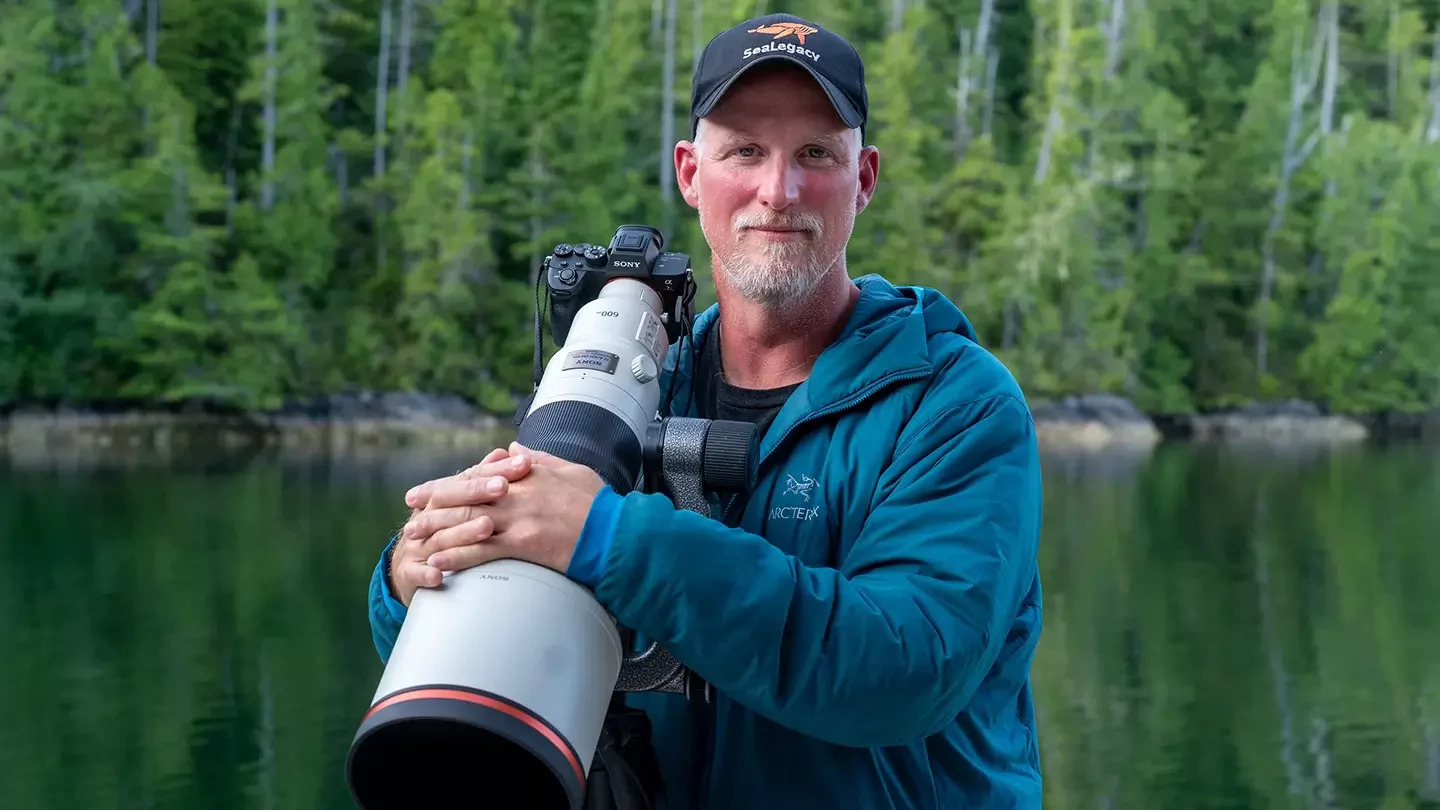 A man wearing a baseball hat that says SeaLegacy and holding a camera with a large lens in front of a body of water surrounded by trees.