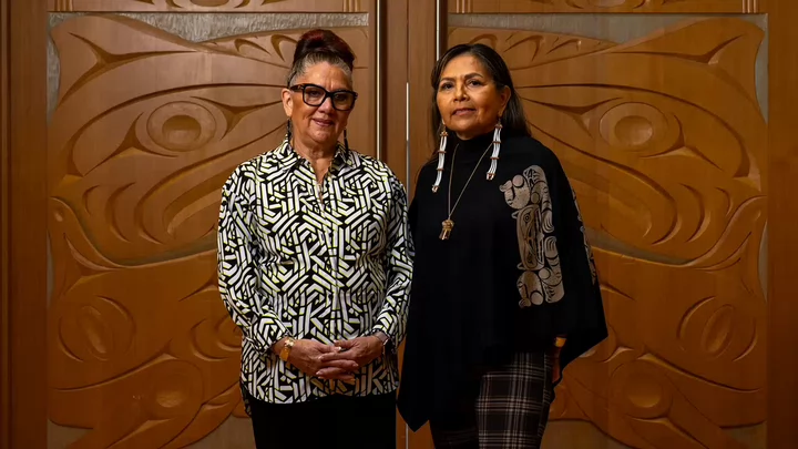 Two women standing side by side in front of a wooden door carved with Indigenous designs. 