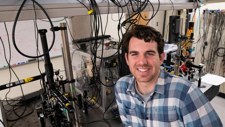 Man with blue plaid shirt smiling in front of complex machinery.