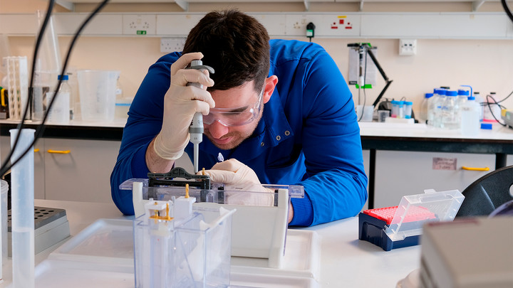 Man in a blue shirt with safety goggles and gloves working in a lab..