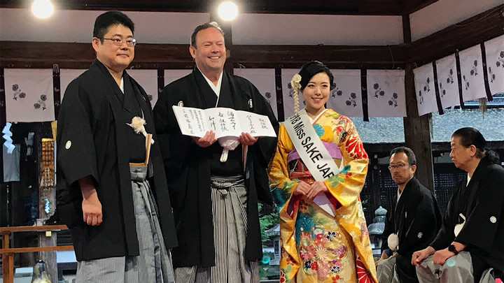 Man in Japanese robe smiling and holding up a certificate with Japanese writing, standing between another man in a robe and a woman wearing a sash that reads Miss Sake Japan.