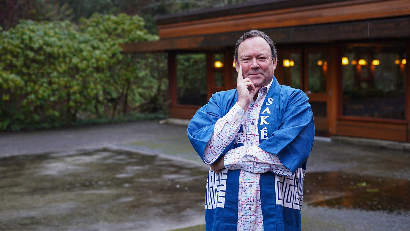 Man wearing a blue Japanese robe that says Sake, standing cross armed with a building and garden in the background.