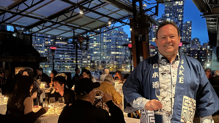 Man in a Japanese robe that reads Sake on the lapel, holding a wine glass and standing on a patio of a busy restaurant at night.