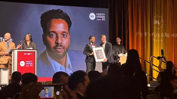 A man with a beard and tuxedo stands on stage and is presented a photo of himself in front of a backdrop that says BIV 40 Under 40 Awards.