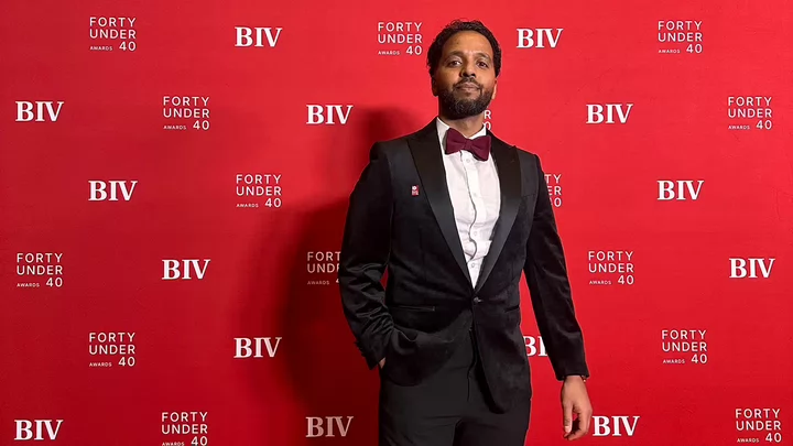 A man with a beard wearing a tuxedo standing in front of a backdrop that reads BIV 40 Under 40.