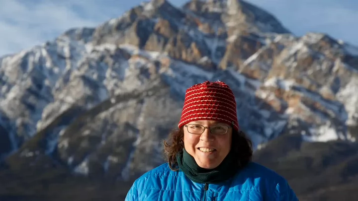 A woman wearing a toque and glasses standing in front of a snow-capped mountain and smiling. 