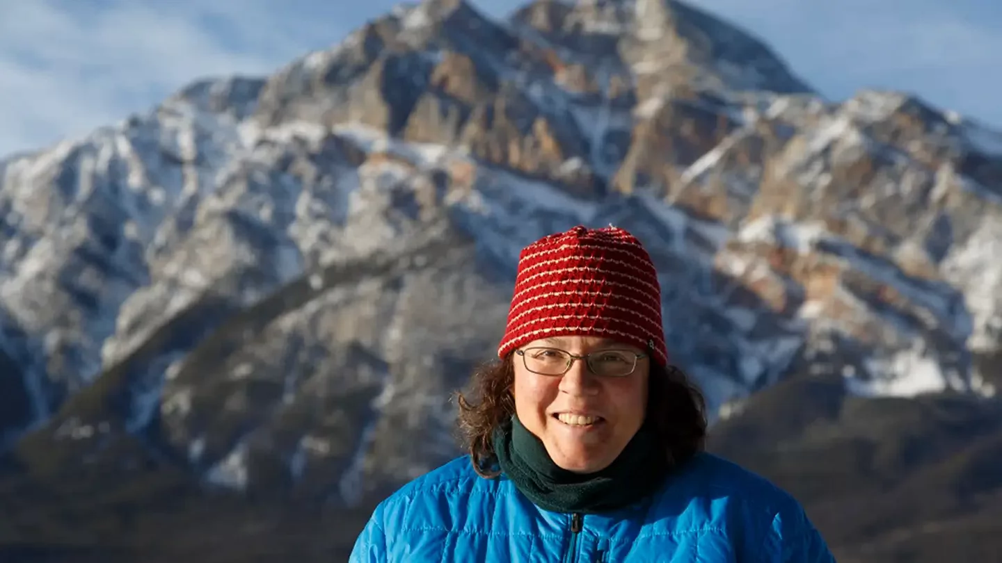 A woman wearing a toque and glasses standing in front of a snow-capped mountain and smiling. 