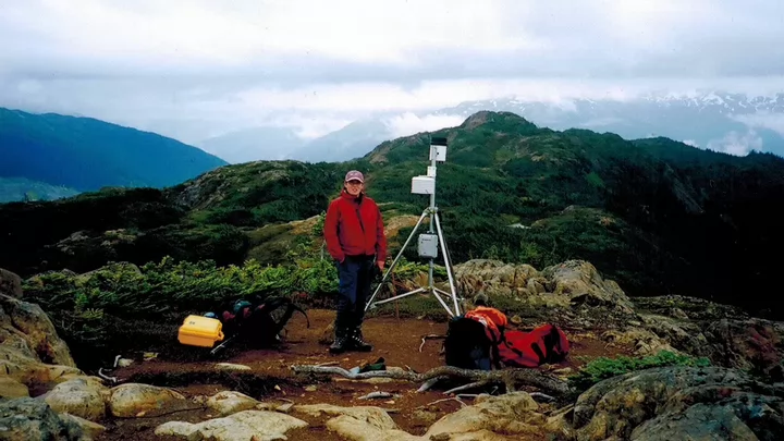 A woman standing atop a mountain beside an assortment of scientific equipment.