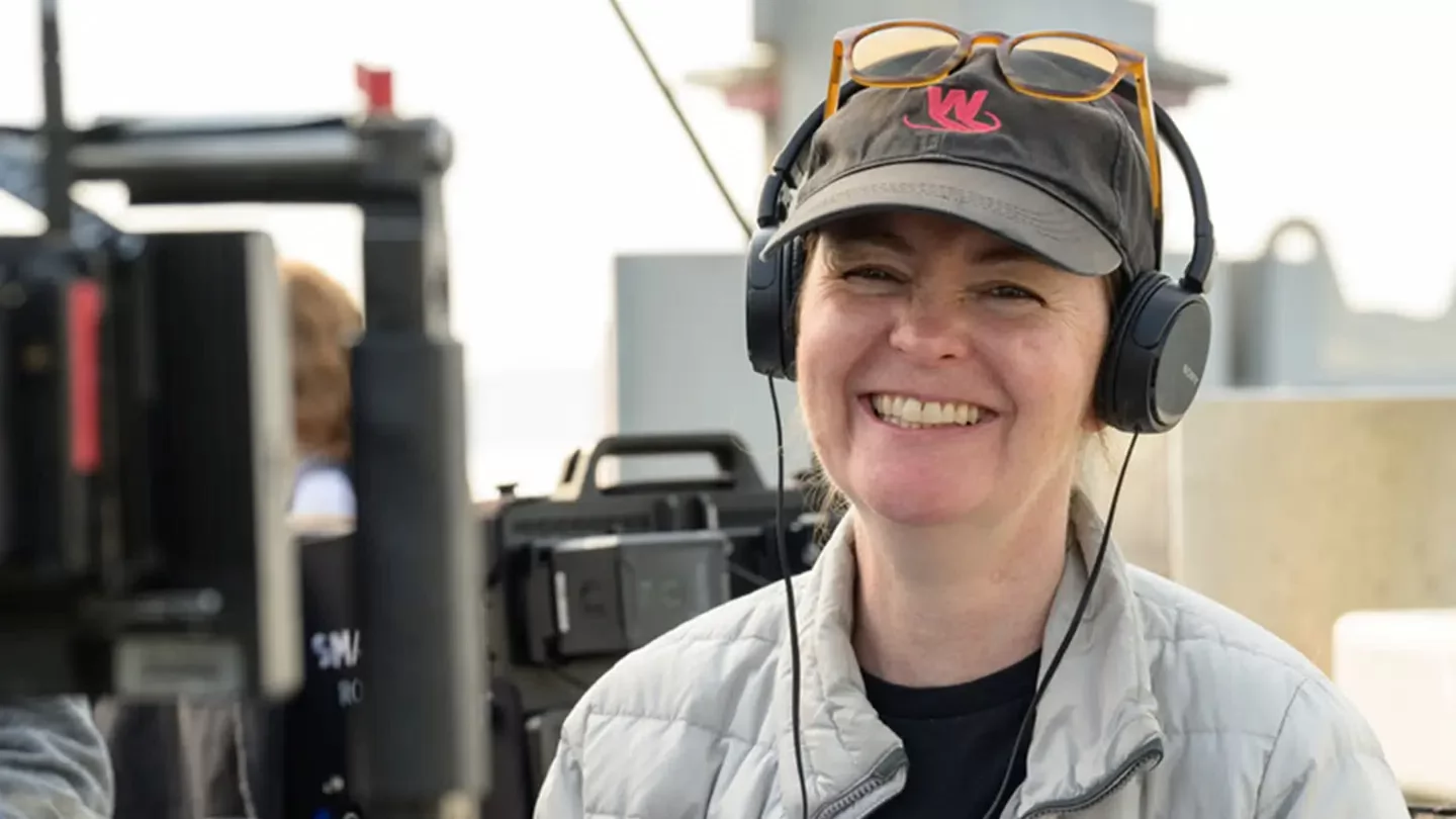 A woman wearing a baseball cap and headphones smiling behind the cameras on a film set. 