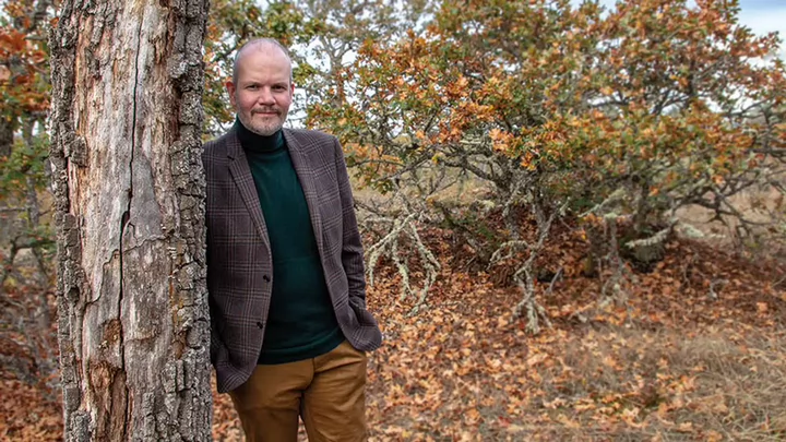 A man in a turtleneck sweater and tweed jacket leaning against a tree. 