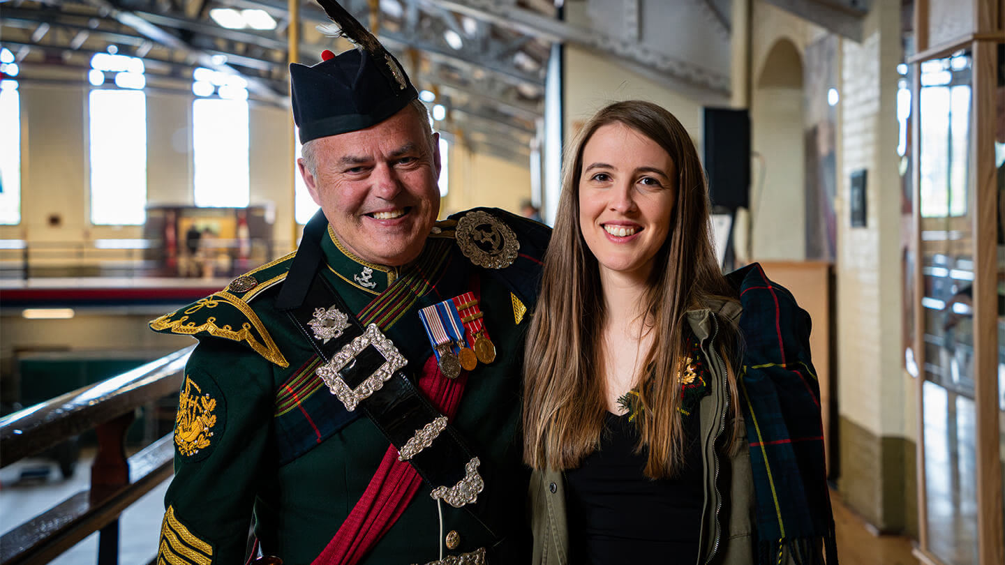 Man wearing a traditional Scottish regiment outfit smiling with a woman wearing a dark green jacket and black shirt underneath.