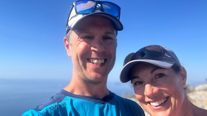 Man with sunglasses atop baseball cap smiling beside woman with sunglasses above baseball cap smiling. 